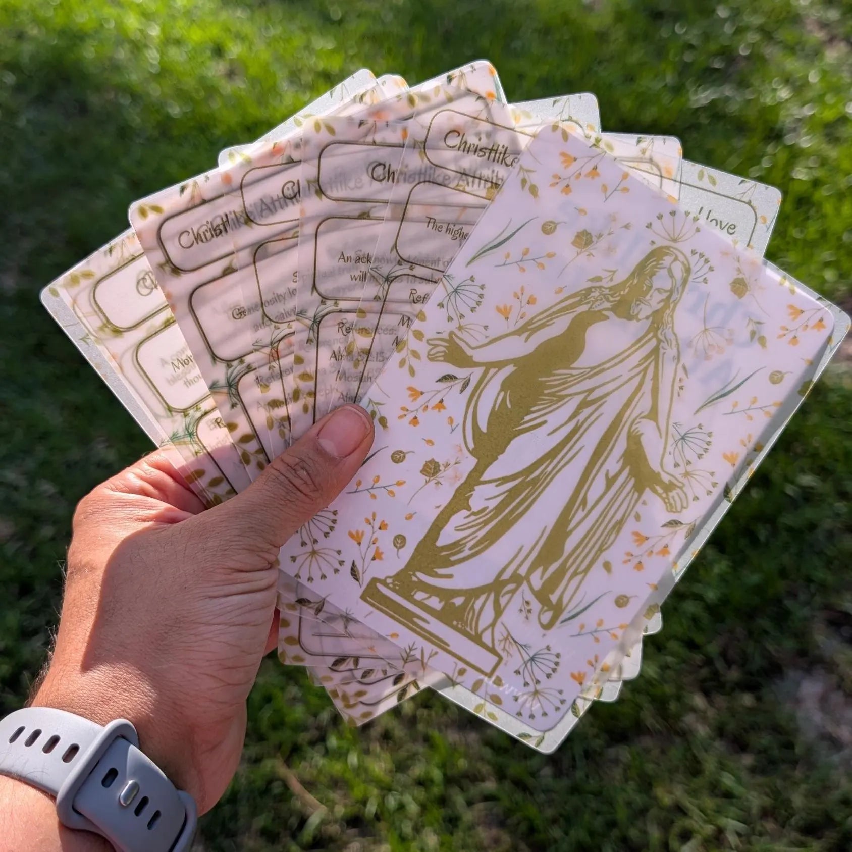 A hand holds a fanned-out deck of religious vellum sheets outdoors on a grassy background. The vellum sheets are translucent with an illustration of the Christ and green text reading 'Christlike attributes.'