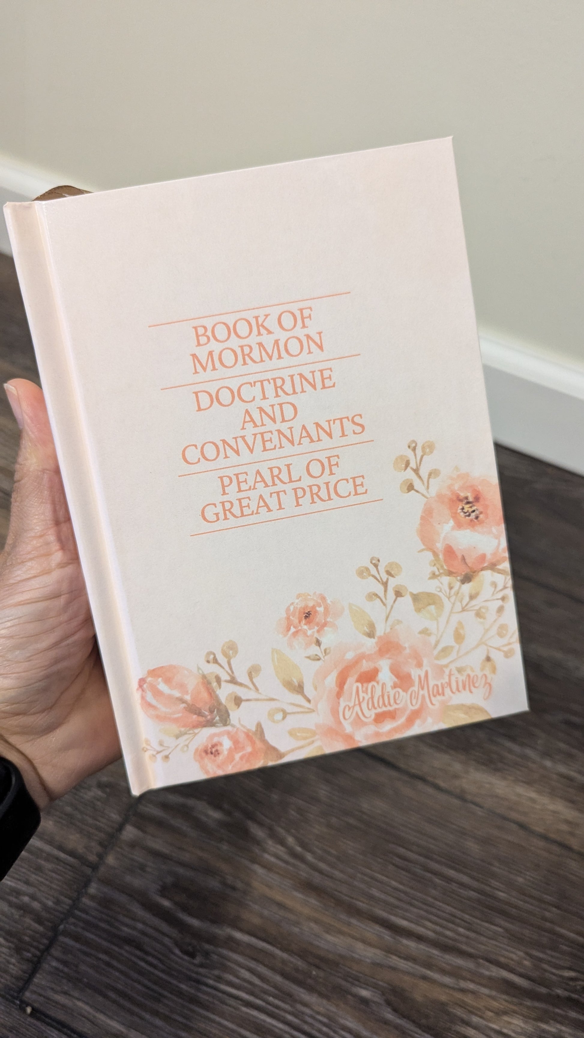 Book of Mormon with floral design held by a hand on a wooden floor.
