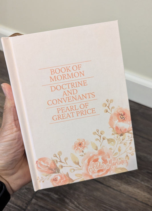 Book of Mormon with floral design held by a hand on a wooden floor.