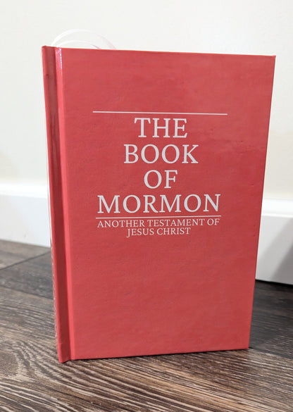 A red hardcover book titled 'The Book of Mormon' standing upright on a dark wood floor against a light wall.
