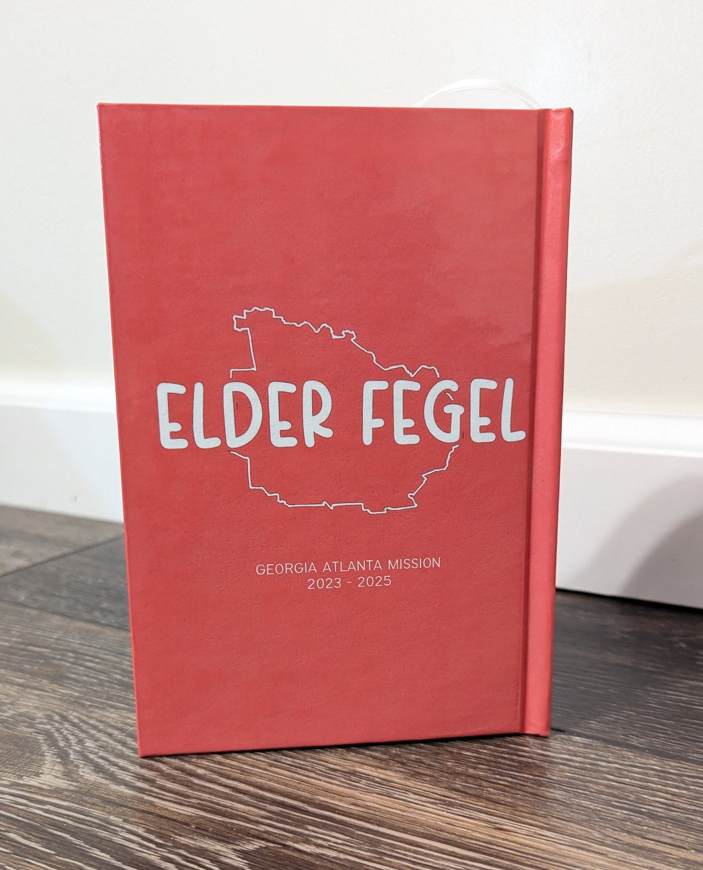 A red hardcover Book of Mormon standing upright on a dark wood floor against a white wall. The cover features a white outline map of Georgia with the text 'ELDER FEGEL' and smaller text reading 'GEORGIA ATLANTA MISSION 2023 - 2025'.