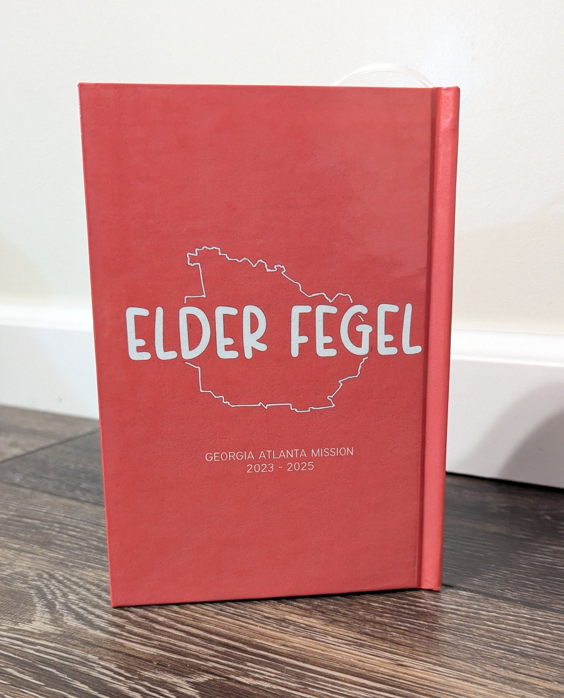 A red hardcover Book of Mormon standing upright on a dark wood floor against a white wall. The cover features a white outline map of Georgia with the text 'ELDER FEGEL' and smaller text reading 'GEORGIA ATLANTA MISSION 2023 - 2025'.