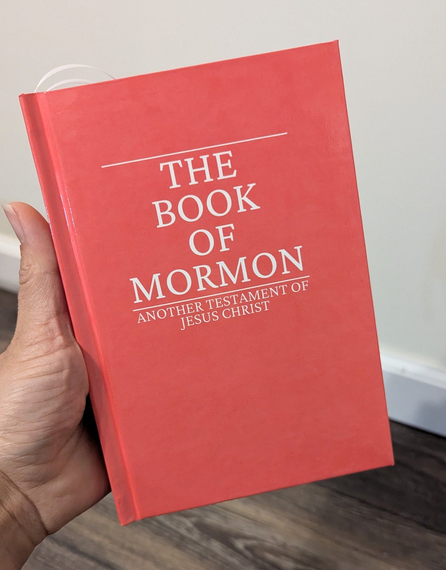 A hand holding a red hardcover book titled 'The Book of Mormon' with white text, set against an indoor background with a light wall and wood floor.