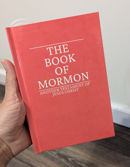 A hand holding a red hardcover book titled 'The Book of Mormon' with white text, set against an indoor background with a light wall and wood floor.