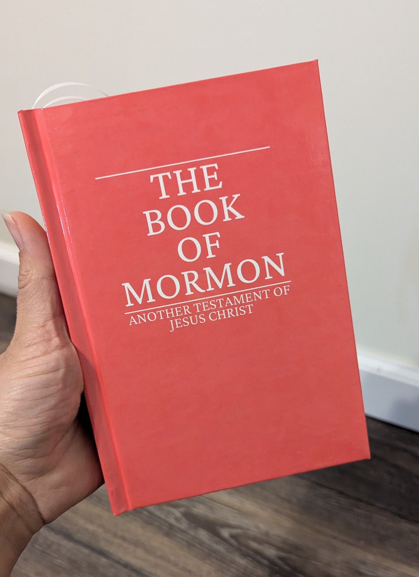 A hand holding a red hardcover book titled 'The Book of Mormon' with white text, set against an indoor background with a light wall and wood floor.