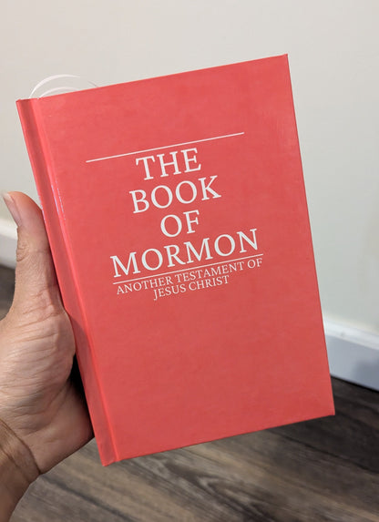 A hand holding a red hardcover book titled 'The Book of Mormon' with white text, set against an indoor background with a light wall and wood floor.