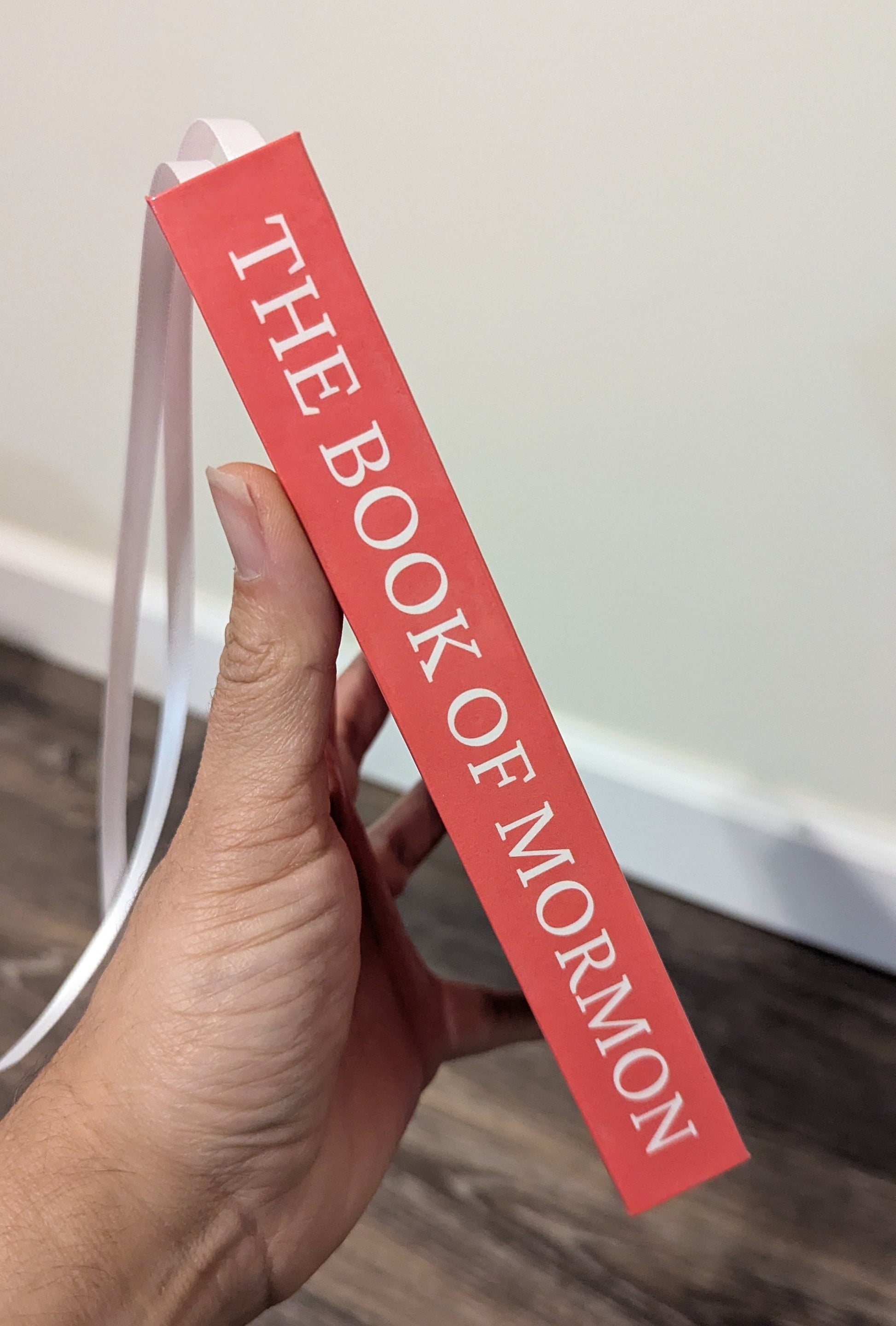 A hand holding a coral-pink book spine prop labeled 'THE BOOK OF MORMON' in white text, with a white ribbon bookmark hanging from the top, against an indoor wall and wood floor background.