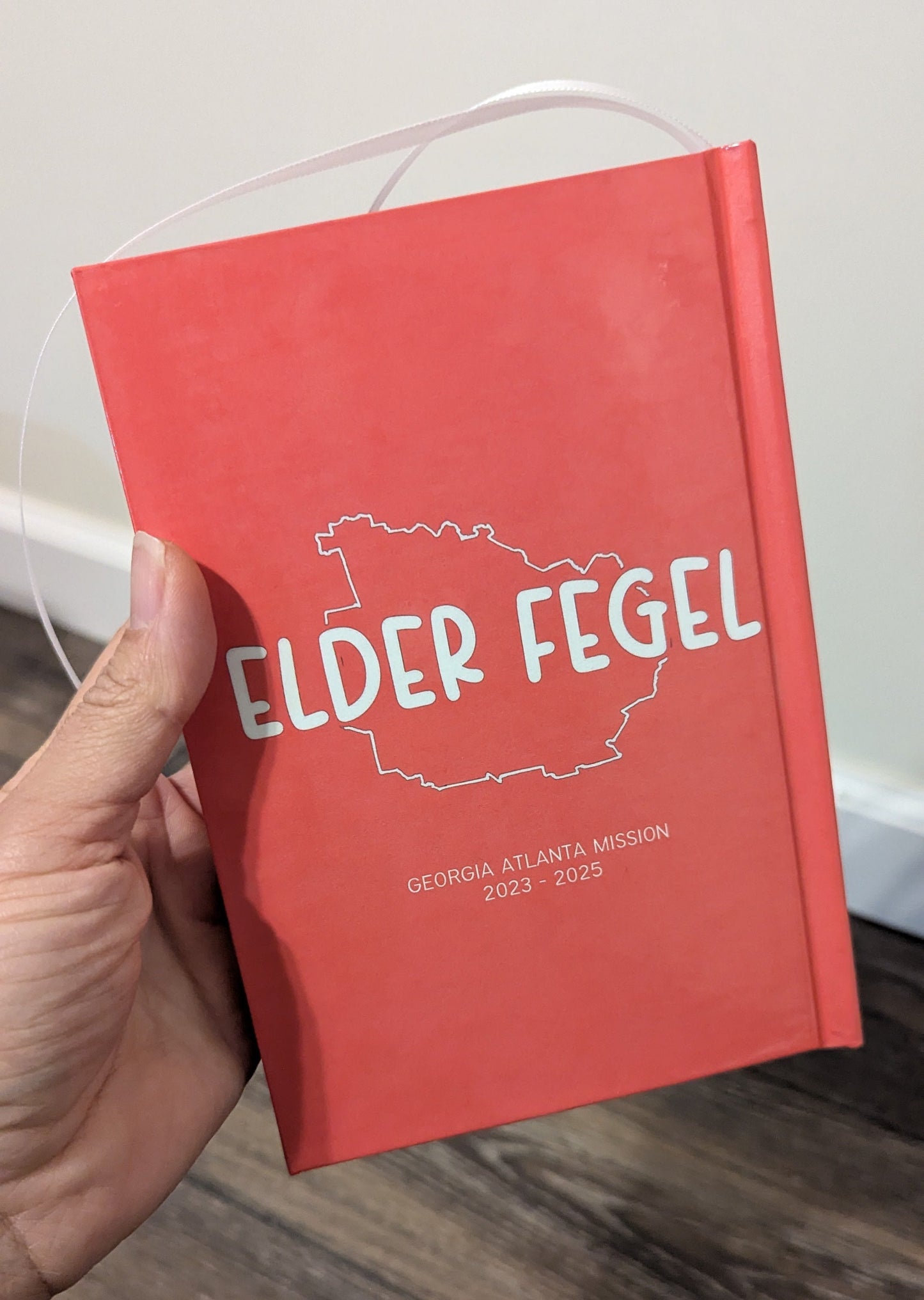 A hand holding a red hardcover notebook or journal with a white printed outline of Georgia and the text 'ELDER FEGEL' on the cover, photographed indoors over a wood floor and light wall.