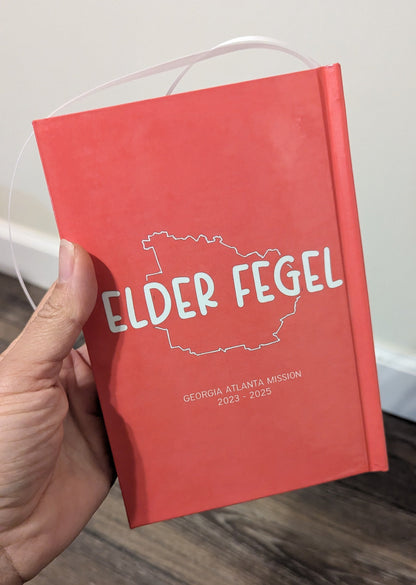 A hand holding a red hardcover notebook or journal with a white printed outline of Georgia and the text 'ELDER FEGEL' on the cover, photographed indoors over a wood floor and light wall.