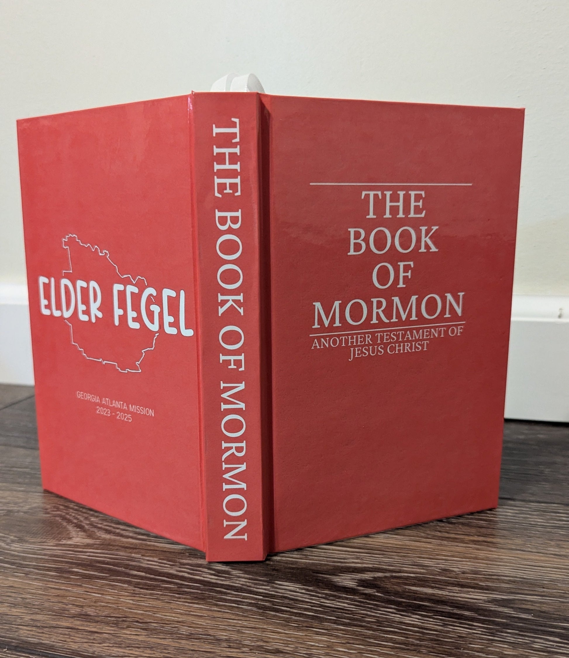 A red hardcover book titled 'The Book of Mormon' by Elder Fegel, standing upright on a dark wood surface against a plain light background.