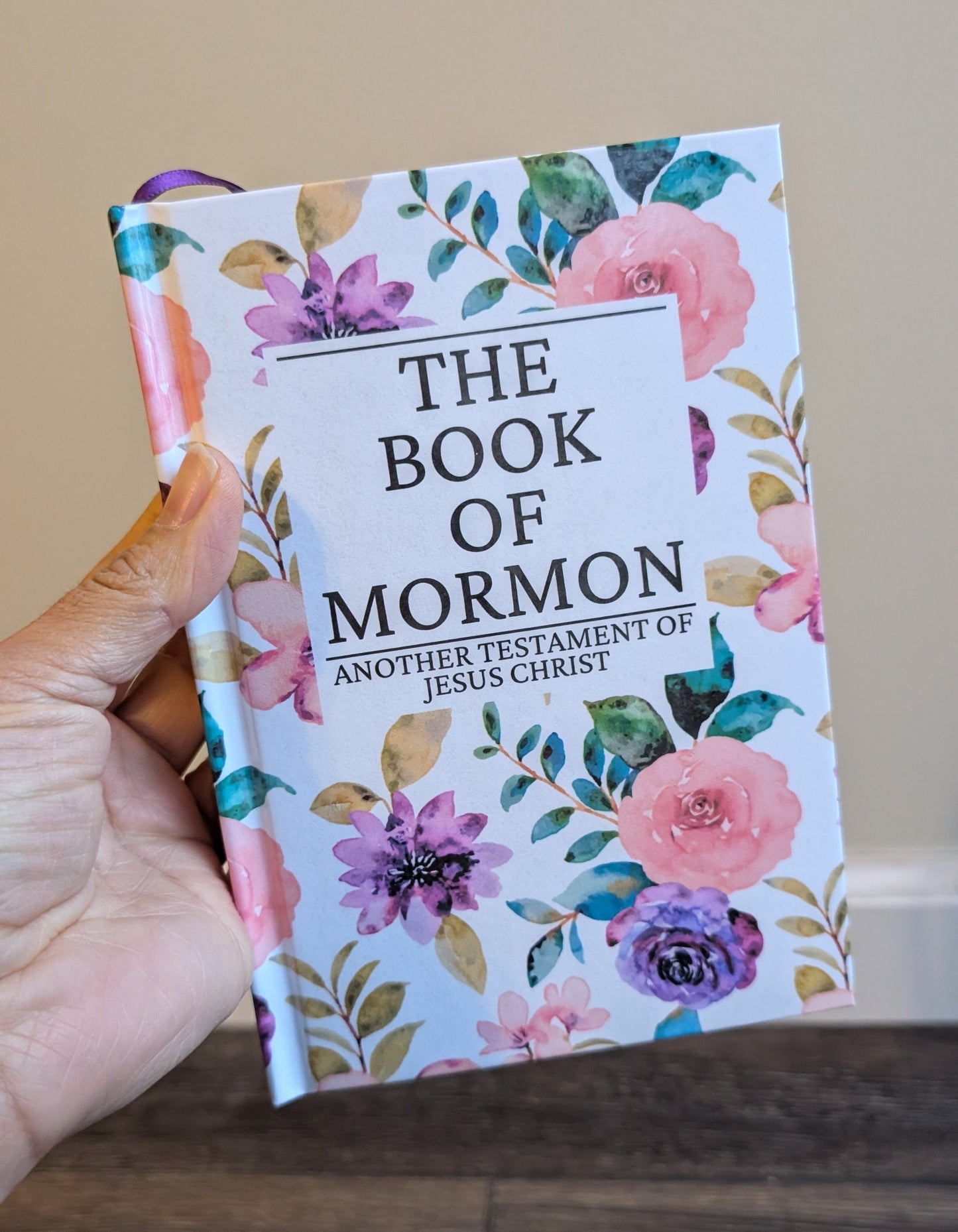 Hand holding 'The Book of Mormon' with floral design on a wooden surface