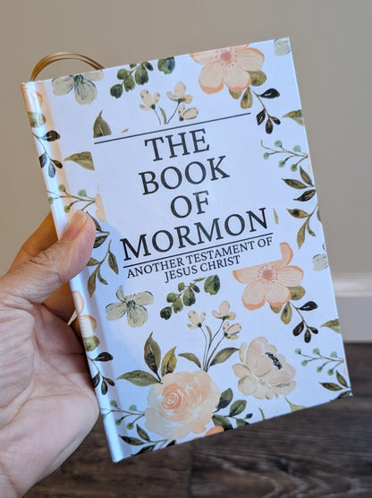Floral decorated 'The Book of Mormon' held by a hand on a wooden surface.