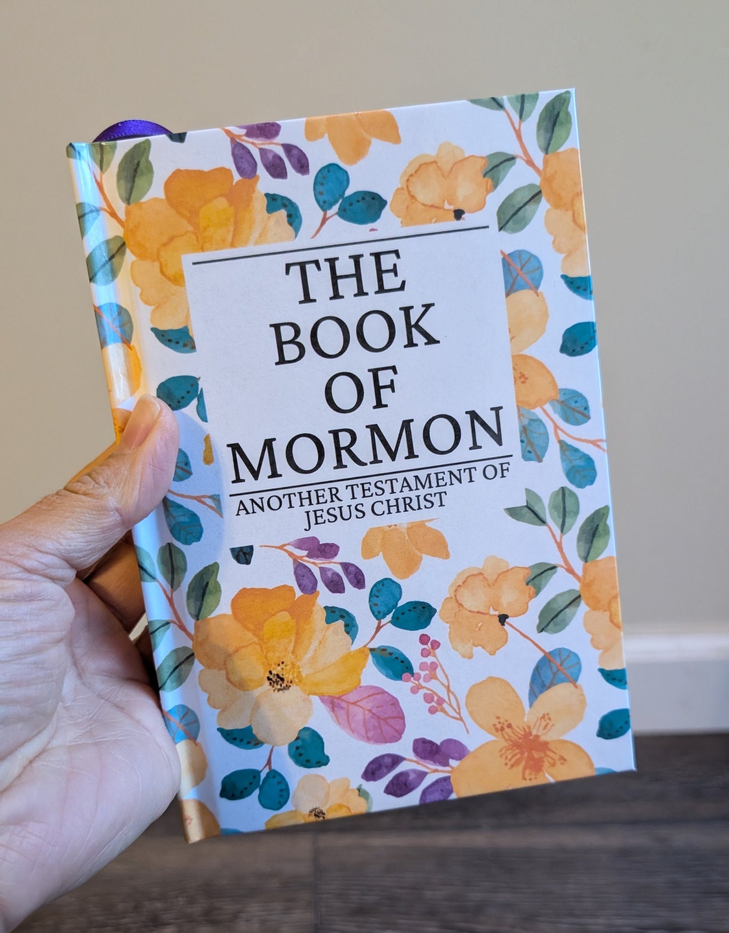 Floral-patterned 'The Book of Mormon' held by a hand on a wooden surface.