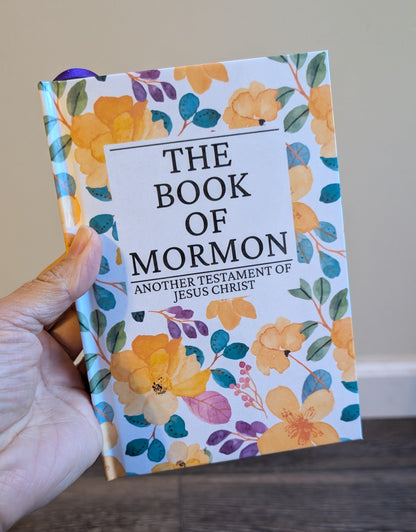 Floral-patterned 'The Book of Mormon' held by a hand on a wooden surface.