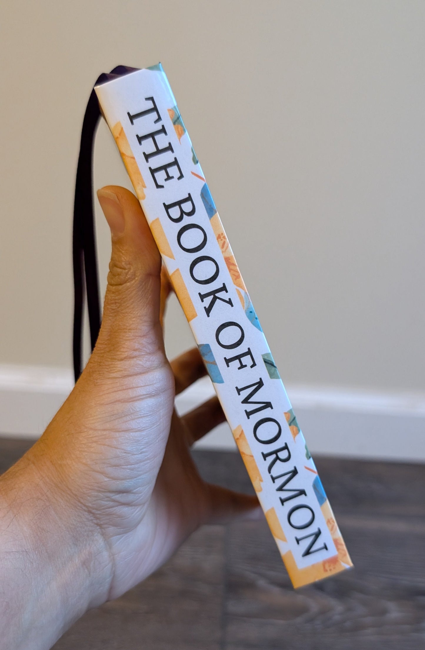 Hand holding a 'The Book of Mormon' book with a strap against a neutral background