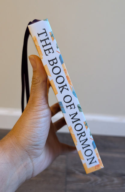 Hand holding a 'The Book of Mormon' book with a strap against a neutral background