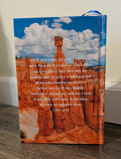 Book of Mormon on a wooden surface. The background of the  Book of Mormon shows the grand canyon. 