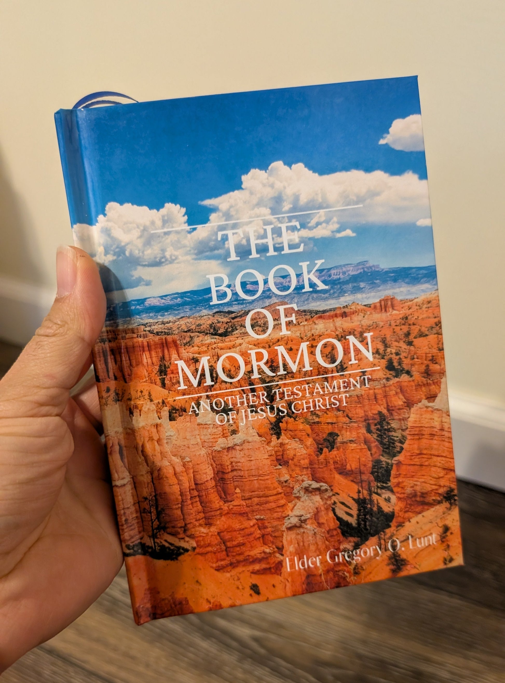 Book of Mormon held by a hand on a wooden surface. The background of the  Book of Mormon shows the grand canyon. 
