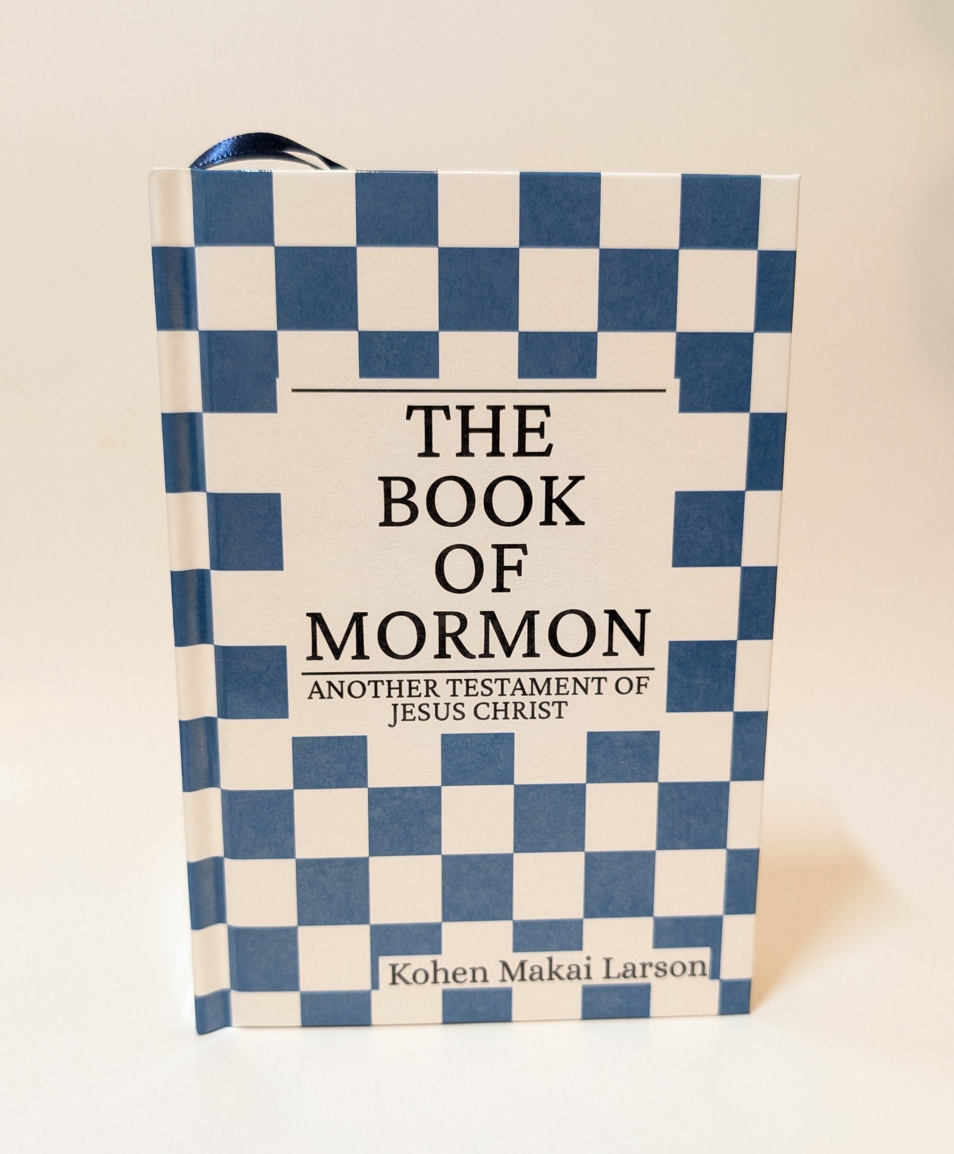 A hardcover book titled 'The Book of Mormon', featuring a blue and white checkered pattern on the cover with a white central panel and black text.