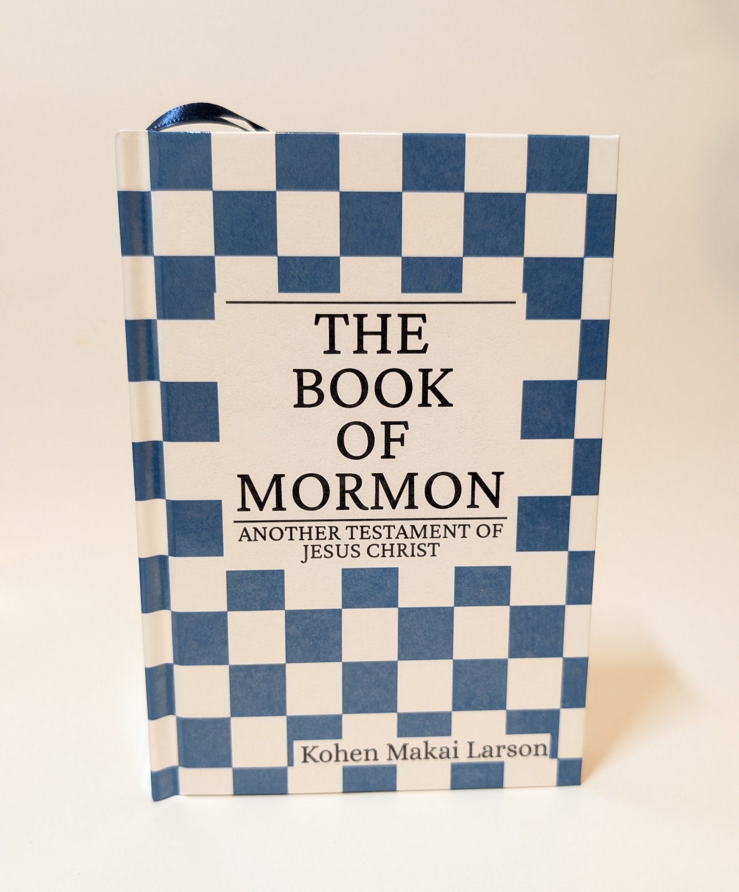 A hardcover book titled 'The Book of Mormon', featuring a blue and white checkered pattern on the cover with a white central panel and black text.
