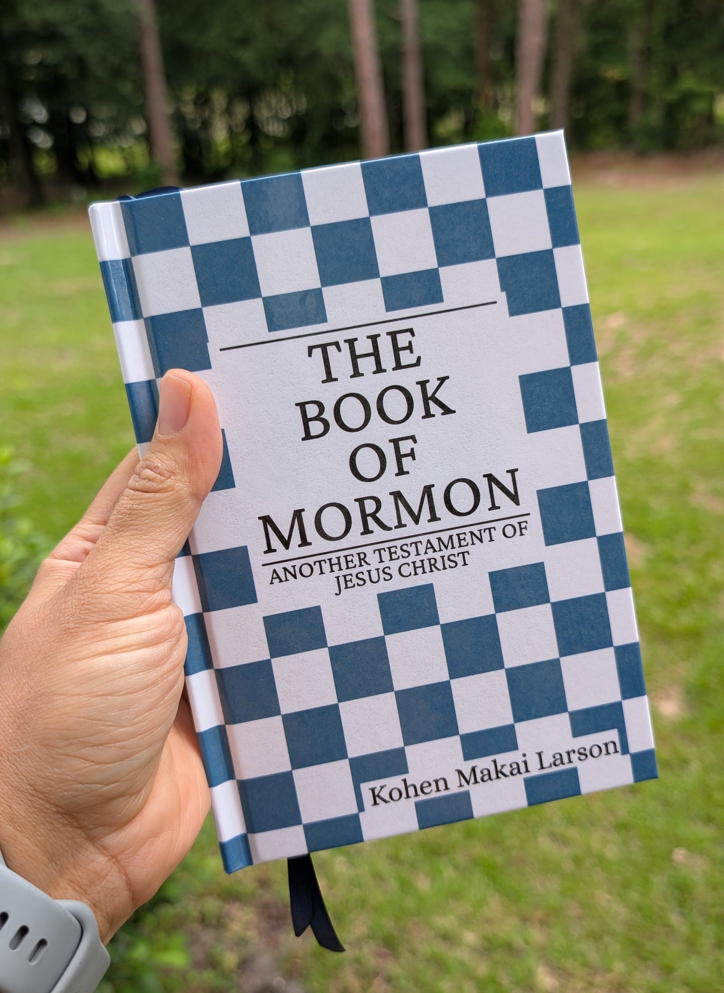 A hand holding a hardcover book titled 'The Book of Mormon' against an outdoor background of grass and trees. The book cover has a blue and white checkered pattern with black text.