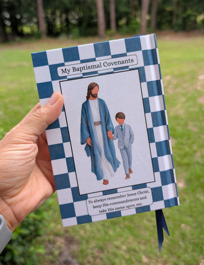 A hand holds a small printed booklet outdoors in a grassy wooded area. The booklet has a blue-and-white checkered border and a central illustration of Jesus and a child, with the title 'My Baptismal Covenants' visible at the top.