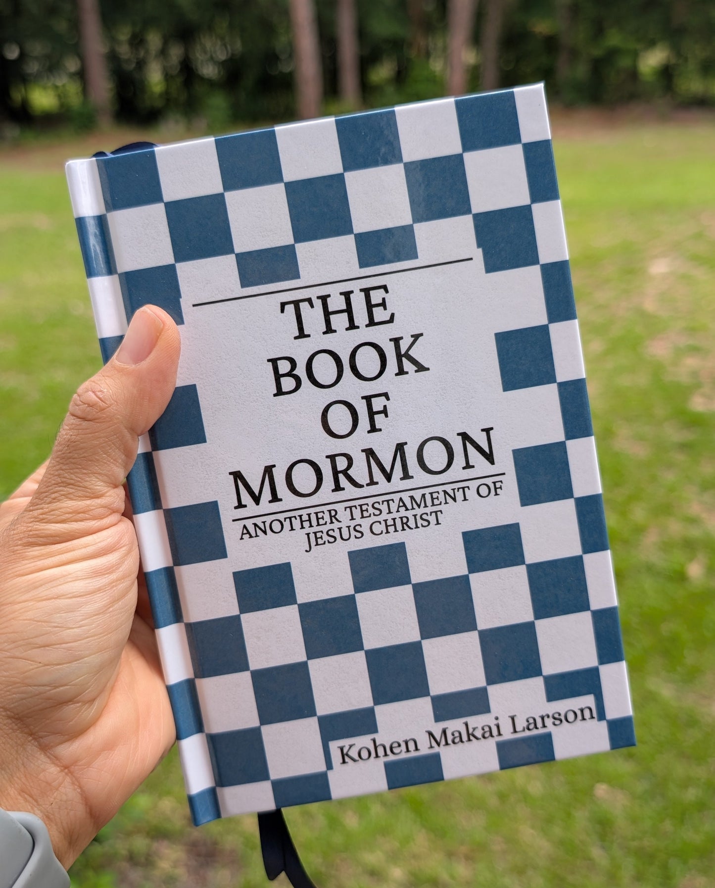 A hand holding a hardcover book titled "The Book of Mormon" outdoors, with a blue-and-white checkered cover and blurred trees and grass in the background.