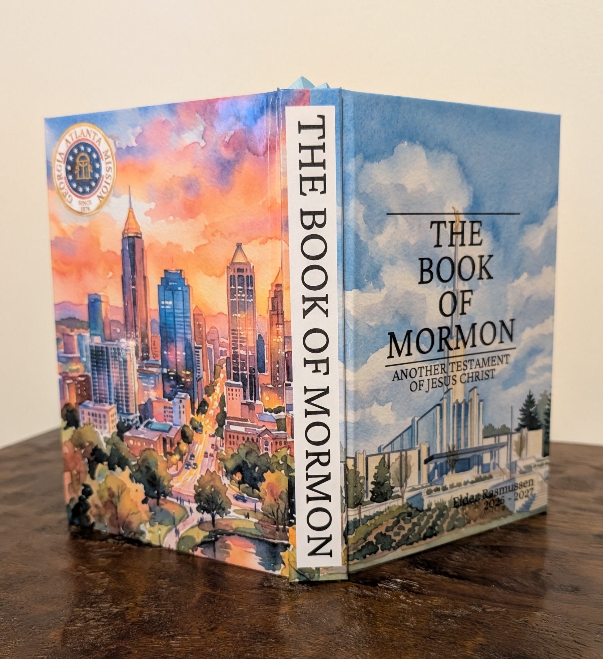 The Book of Mormon hardcover Bible standing upright on a dark wooden table against a plain beige wall. The cover features a colorful cityscape illustration on the front and spine, with the title 'THE BOOK OF MORMON' clearly visible.