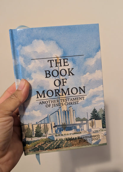 A hand holding a hardcover book titled 'The Book of Mormon' against a plain light wall. The cover features a blue sky and clouds with an illustration of a temple.