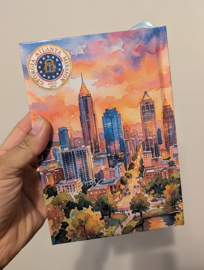 A hand holding a Book of Mormon (showing the back cover) featuring a colorful city skyline illustration against a plain indoor wall.