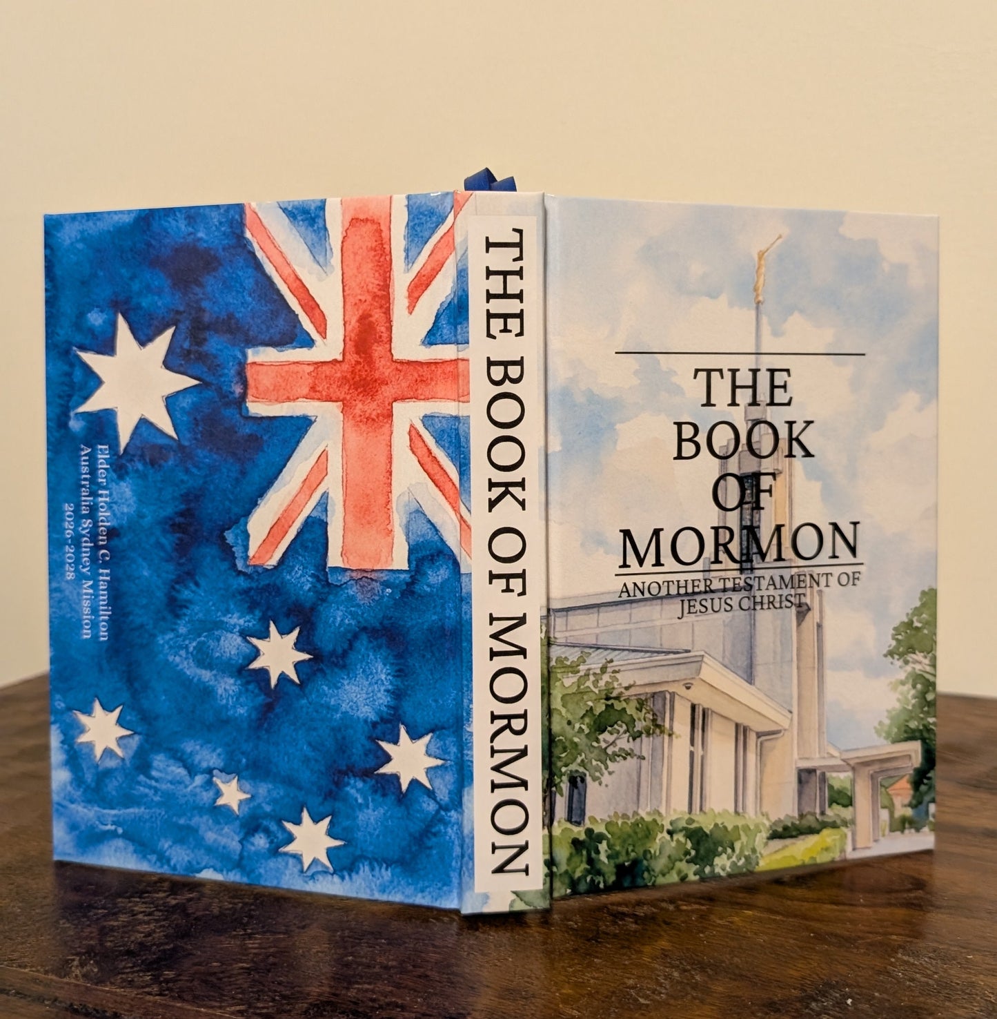 The Book of Mormon, shown open to display its front cover, spine, and inner page. The cover features a watercolor Australian flag on the left and a temple illustration on the right, resting on a dark wooden table against a beige wall.