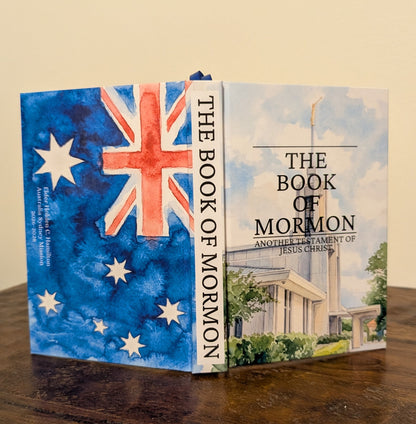 The Book of Mormon standing upright on a wooden table, showing the Australian flag on the front cover and the temple illustration on the spine in watercolor.