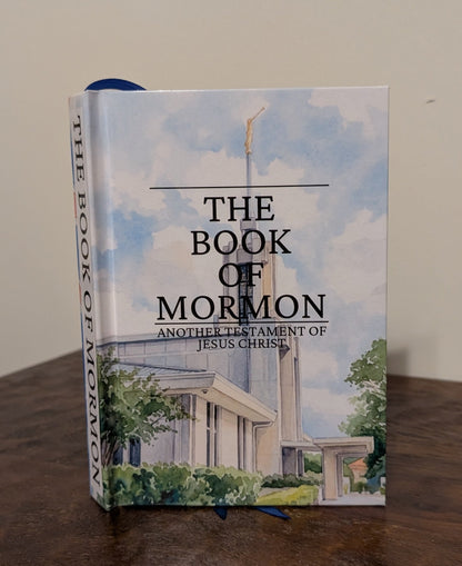 A hardcover book titled "The Book of Mormon" standing upright on a dark wooden table against a plain light wall.