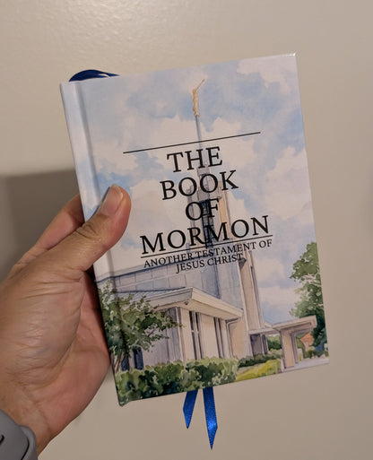 A hand holding a hardcover book titled 'The Book of Mormon' against a plain beige wall. The cover features a light blue sky and temple illustration, with blue ribbon bookmarks visible.