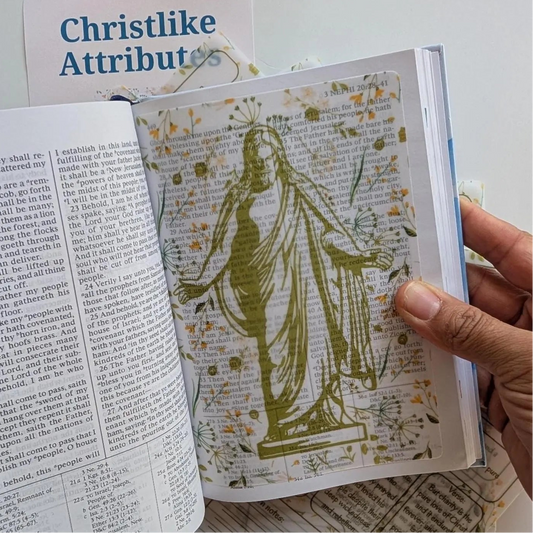A hand holds an open book of Mormon spread showing a vellum sheet with an image of Christ.
