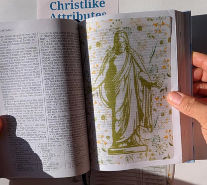 An open book with a vellum sheet inside of it. The vellum sheet depicts a picture of Christ with His hands and arms extended.