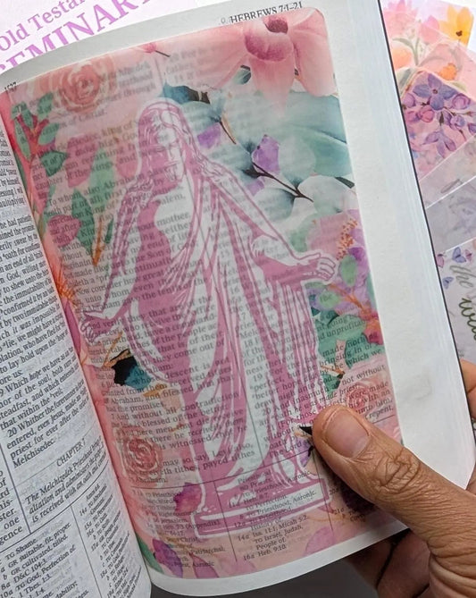A hand holds an open book or journal page featuring a pink floral and religious illustration made from layered magazine pages, with additional decorated translucent sticker sheets visible on the right.